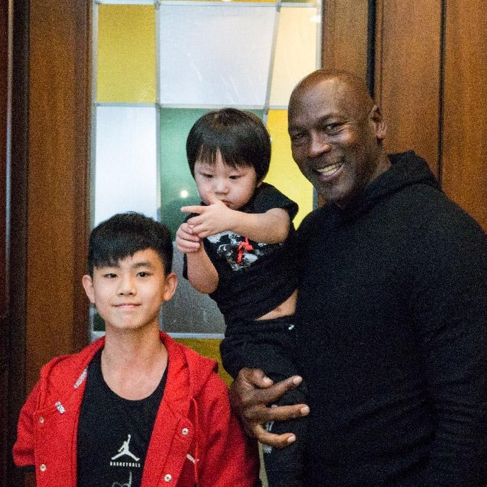 Michael Jordan smiling while holding a young child, standing beside another child in a red jacket